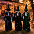 Three witch figures in black robes and pointed hats with small lights holding hands in a yard