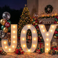 Marquee-style lighted JOY letters with warm bulbs in front of a decorated Christmas tree