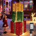 Three stacked lighted gift boxes in red, green and gold with a girl in a Santa dress nearby